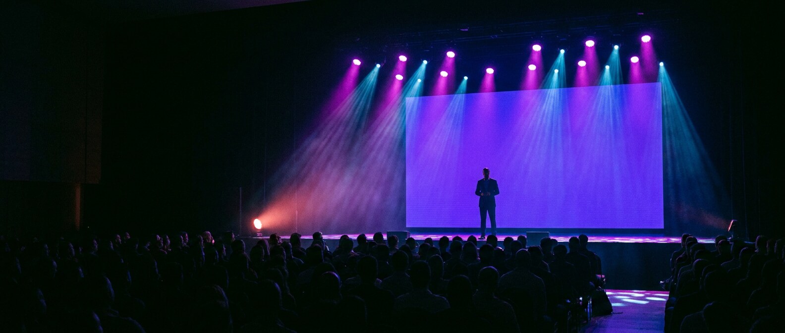 A speaker on stage at the Raise Next summit, lit by violet and cyan stage lighting